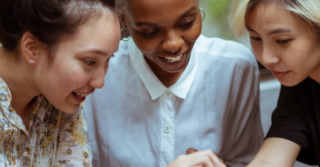 Three diverse women smile while using a smartphone in a cozy indoor setting.