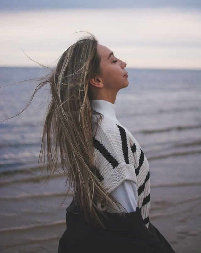A woman with closed eyes embracing the ocean breeze on a beach at sunset, reflecting calmness.